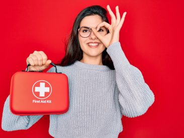 Young beautiful girl holding first aid kit medical box over red background with happy face smiling doing ok sign with hand on eye looking through fingers