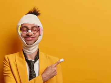 Cheerful man with funny face expression, closes eyes, recovers after injury, has abrasions and bruises, has head concussion, plaster on nose and finger, shows way to chemist, isolated on yellow wall